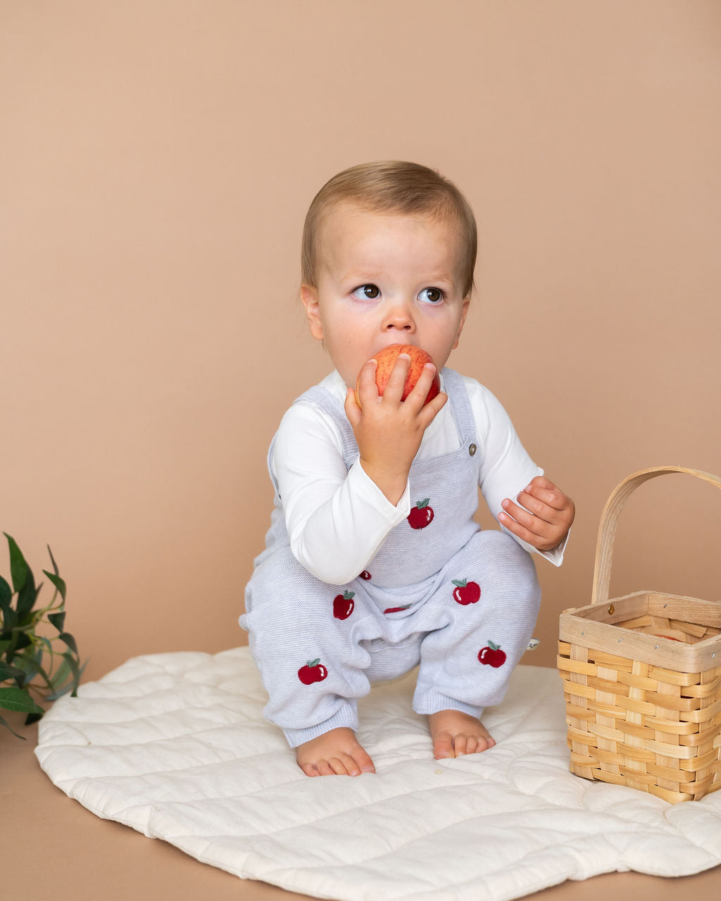 Baby wearing Viverano Organics organic cotton baby overall set with red apple embroidery sitting on quilted mat