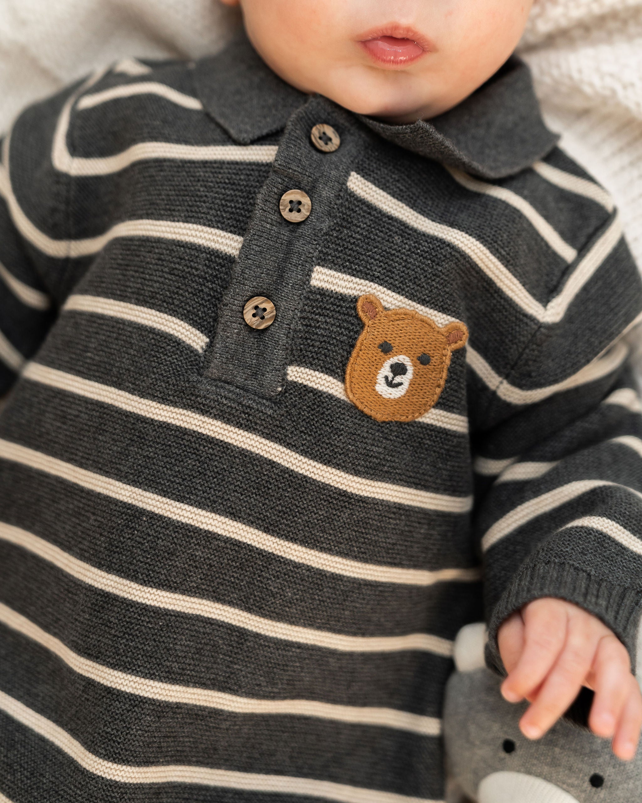 Close-up of a baby wearing a dark gray and cream striped organic knit polo sweater with a brown teddy bear face patch on the chest.