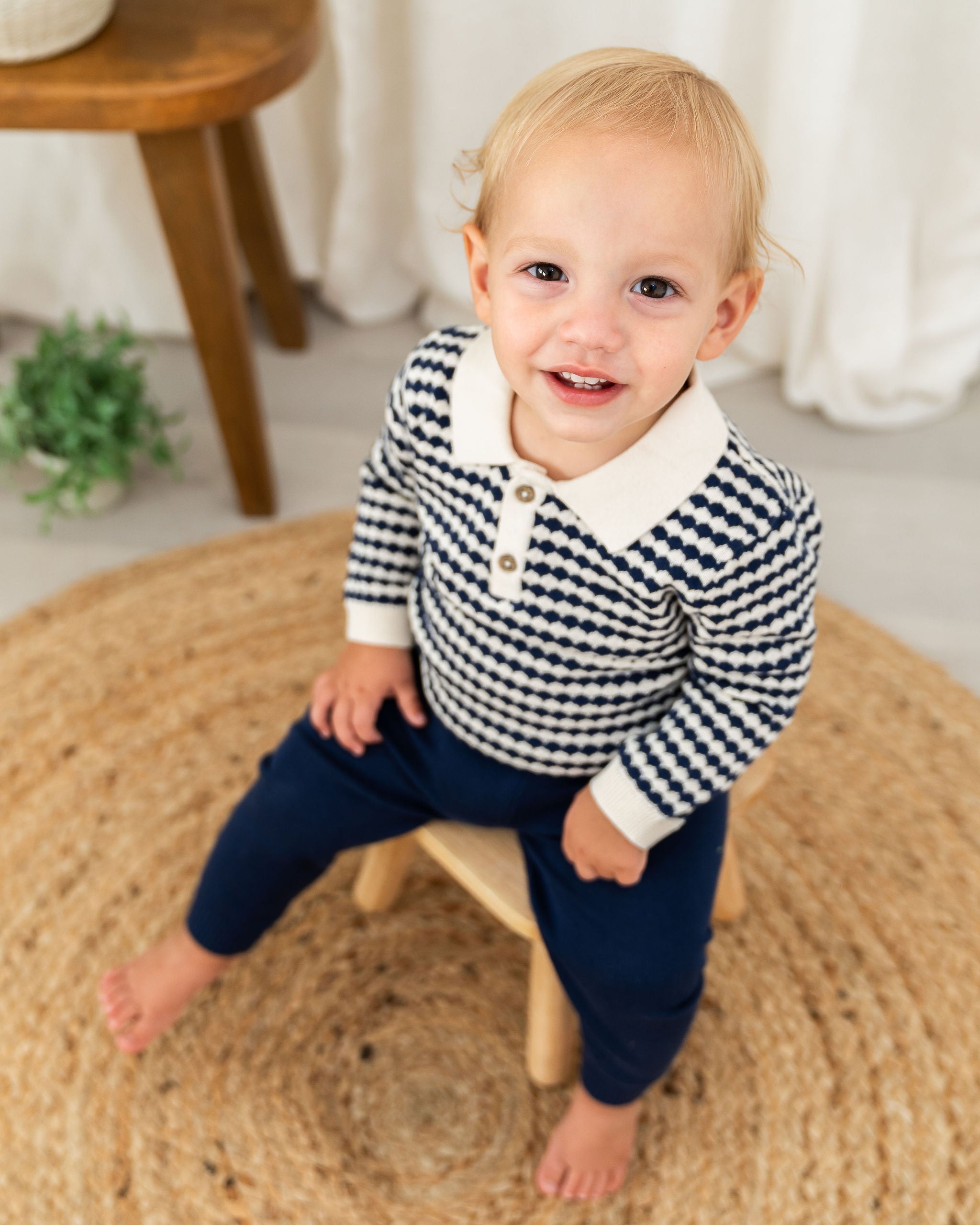 A toddler boy sitting on a small wooden stool, wearing an organic navy and white patterned long-sleeve polo shirt with a white collar and navy pants, in a bright indoor setting.