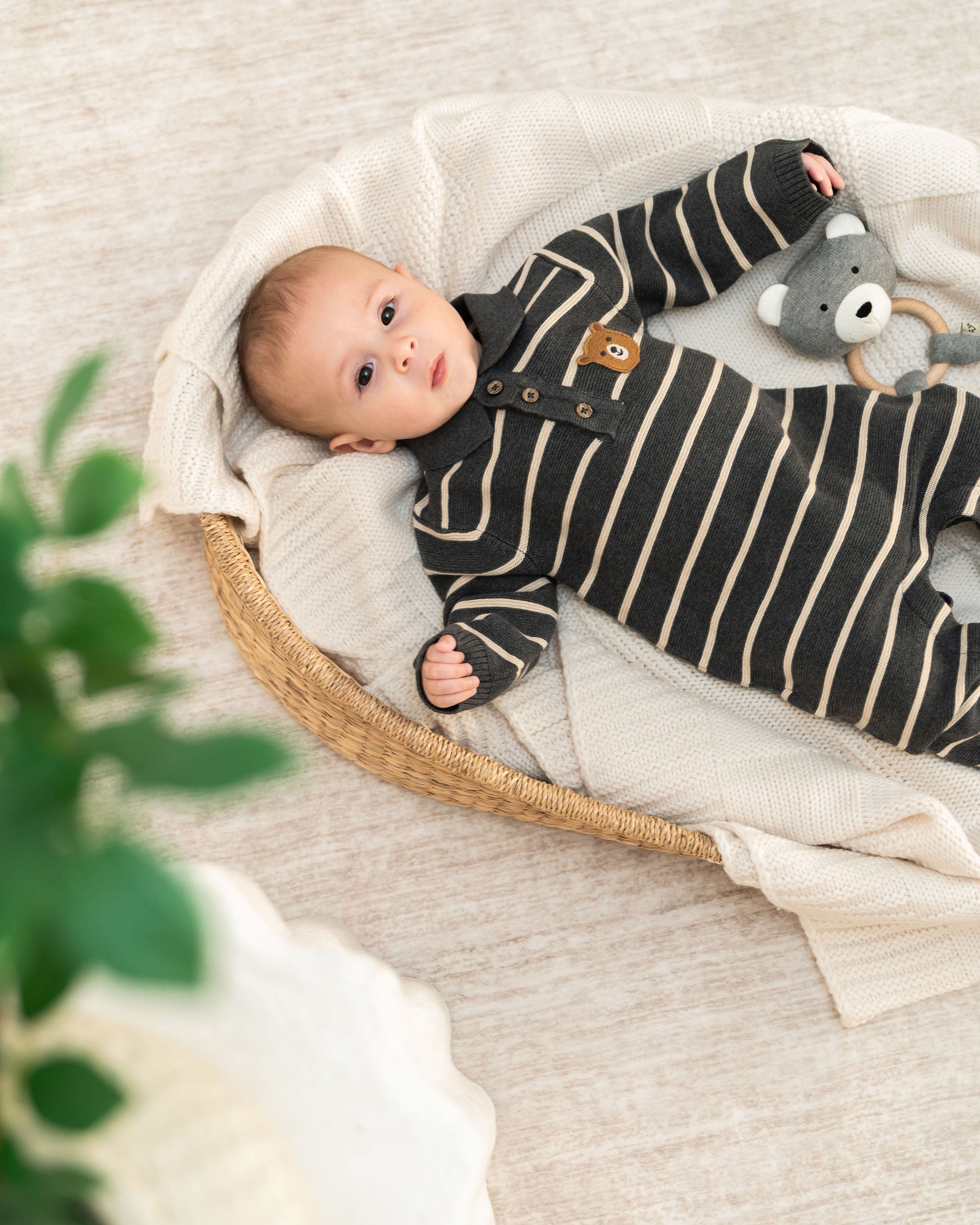 A baby lying on a cream knit blanket in a woven basket, wearing a black and cream striped long-sleeve romper with a small brown bear patch on the chest. A gray bear rattle toy is tucked near the baby.