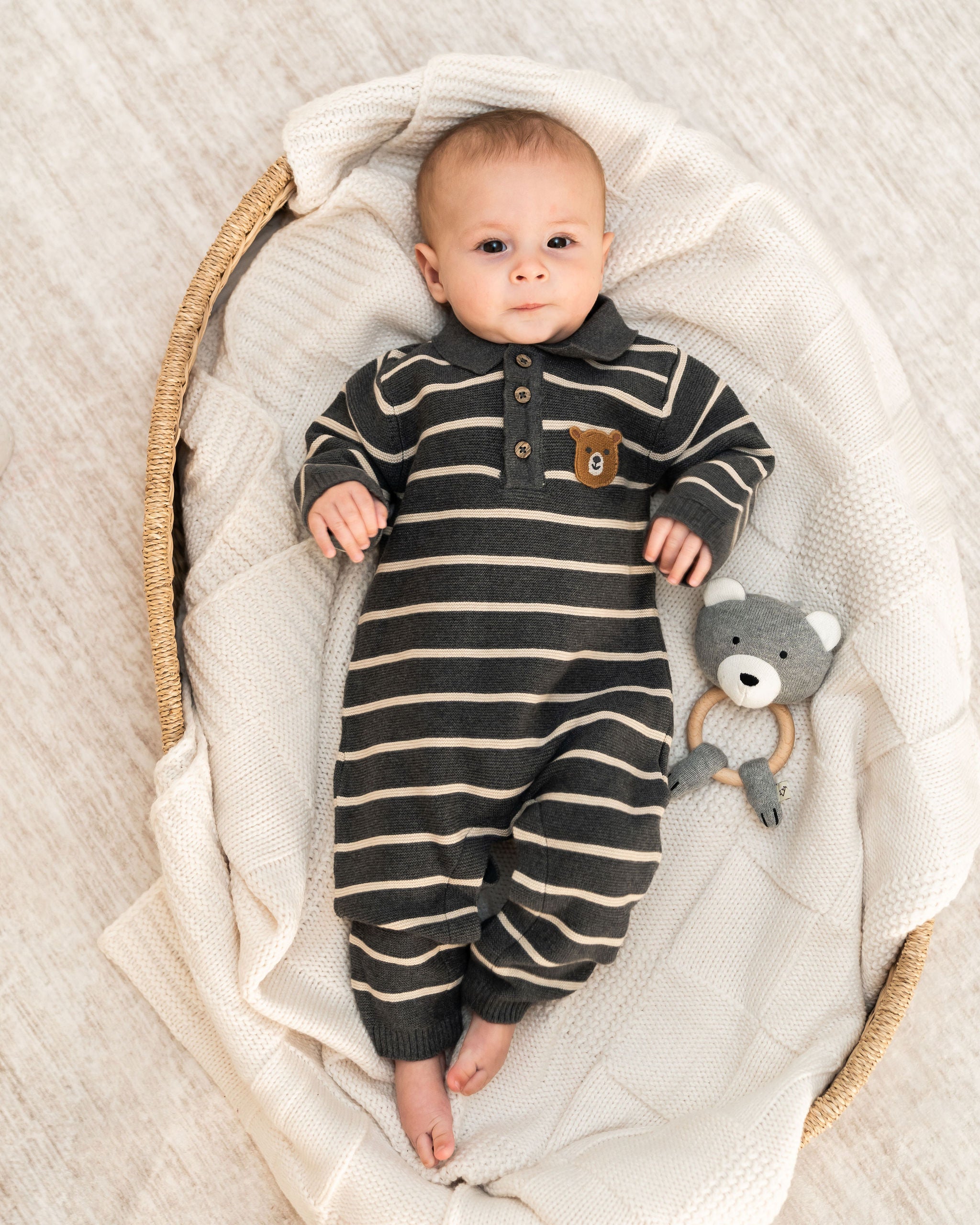 A baby lying on a cream knit blanket inside a woven basket, wearing a dark gray and cream striped long-sleeve organic knit footless onesie with a small brown bear patch on the chest, next to an organic bear rattle toy.