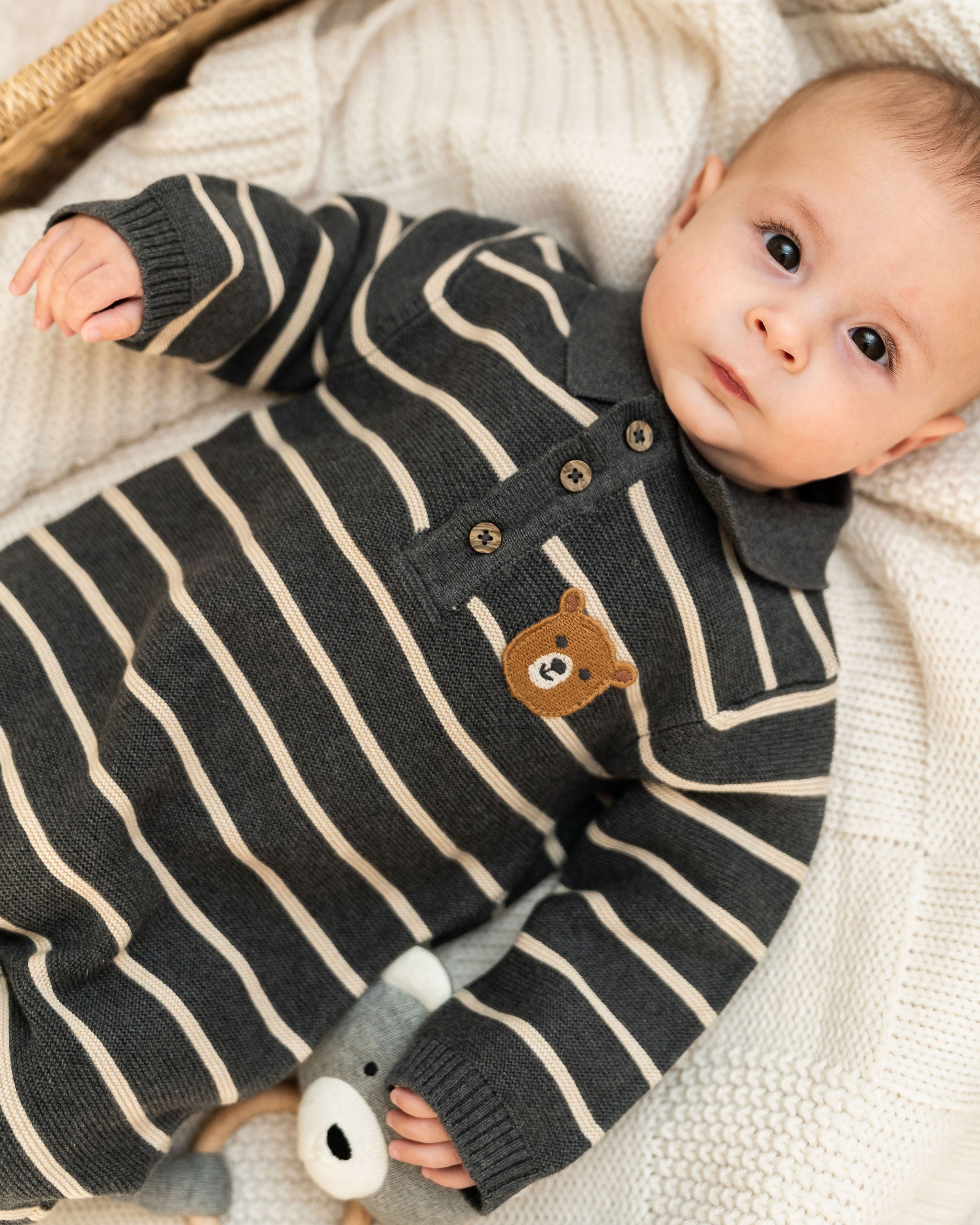 A baby lying on a cream knit blanket wearing a dark gray and cream striped organic knit romper with a small brown bear face patch on the chest.