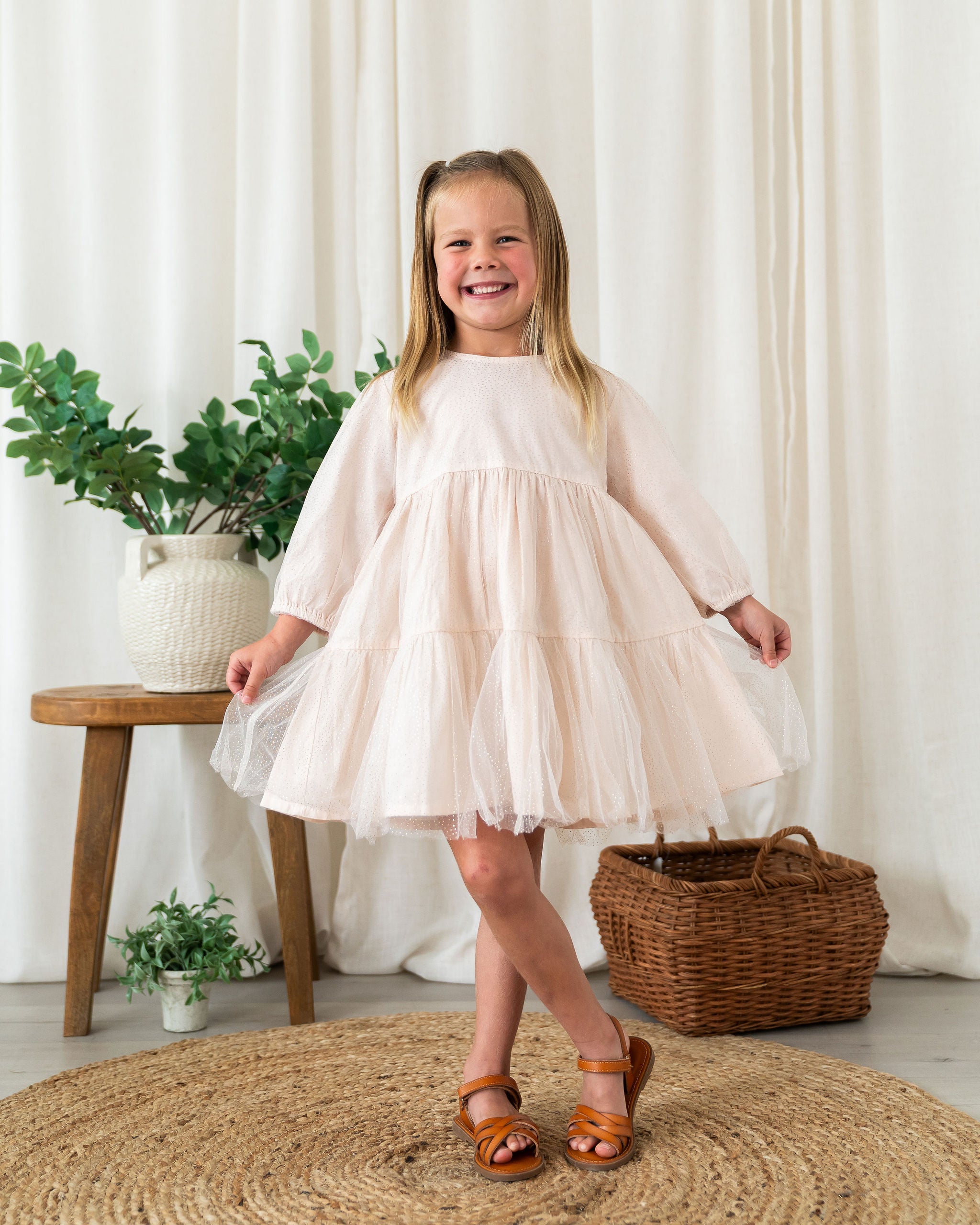 A toddler young girl with blonde pigtails stands smiling in a cream-colored tiered organic tulle dress with three-quarter sleeves, paired with brown leather sandals. She is posing in a styled room with a woven rug, a wicker basket, and a wooden stool with a potted plant against a backdrop of cream curtains.