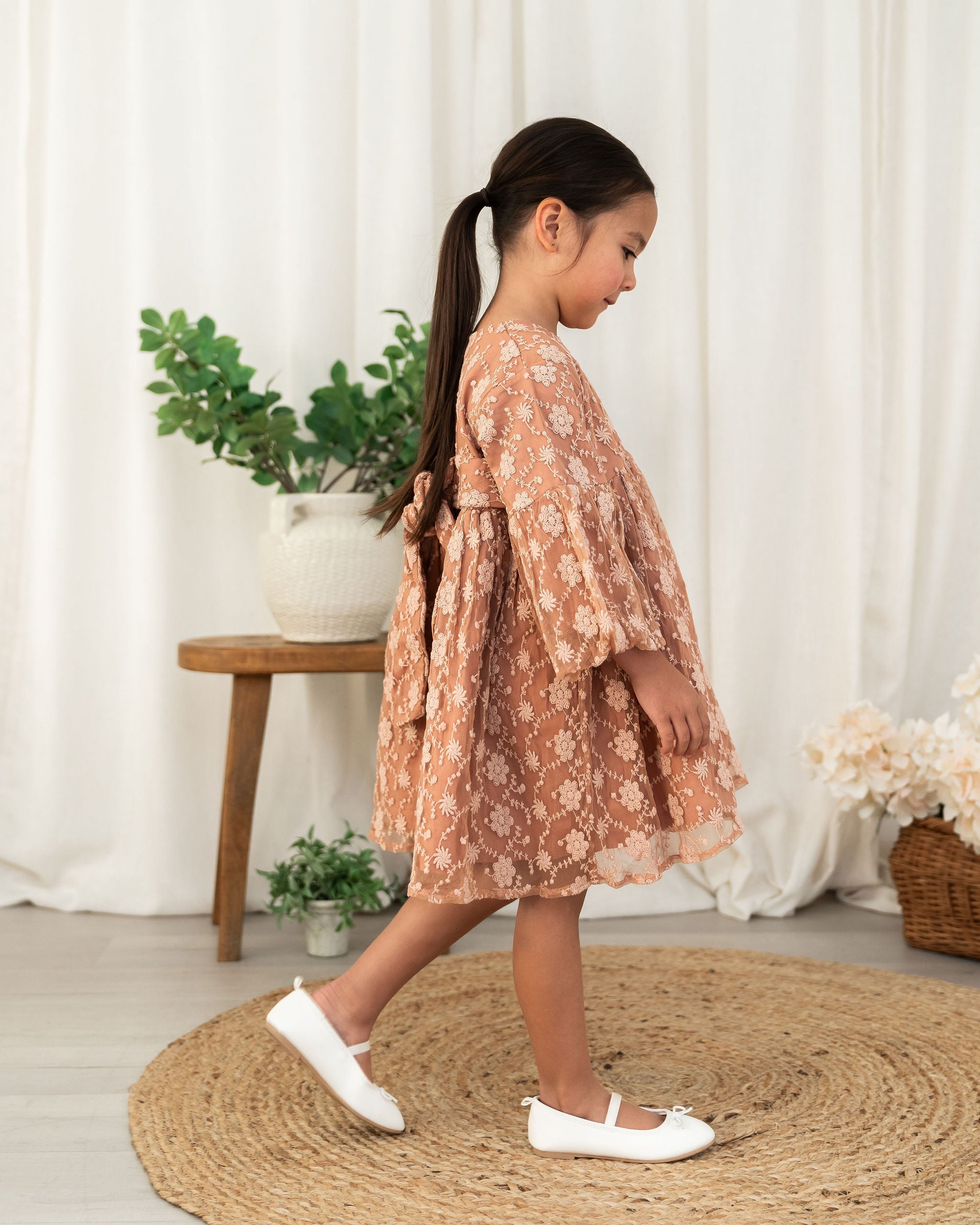 A toddler girl with a dark ponytail is wearing a dusty rose organic lace dress with white floral patterns, walking on a round jute rug in a bright room with white curtains and potted plants.